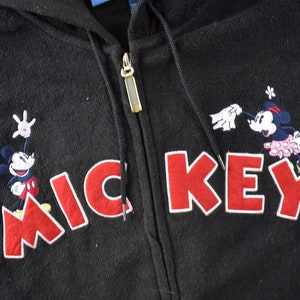May include: Black hooded sweatshirt with a red embroidered Mickey Mouse design. The design features Mickey Mouse and Minnie Mouse with the word "MICKEY" in red.