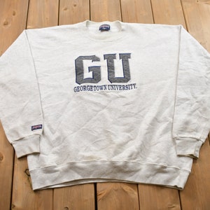 Peut inclure: Un sweat-shirt blanc avec un logo "GU" brodé en gris et le texte "GEORGETOWN UNIVERSITY" en dessous.