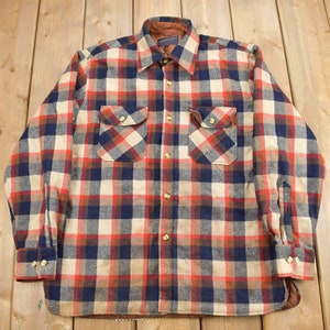 Pode incluir: Uma camisa de flanela de manga comprida com um padrão xadrez vermelho, azul e branco. A camisa tem um fecho de botões na frente e dois bolsos no peito.