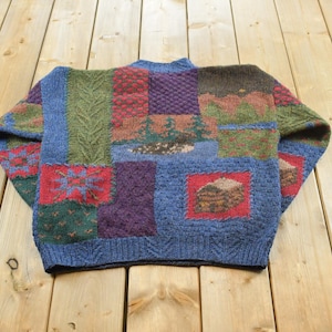 May include: A long-sleeve knit jumper featuring a patchwork design with various textures and colours including blue, green, red, and purple. The pattern includes geometric shapes, cable knit sections, and a landscape scene with trees and a cabin.