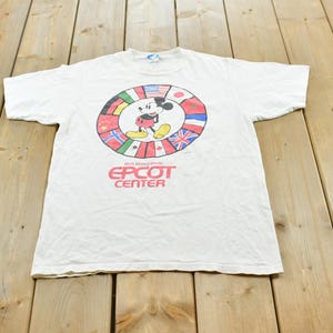 May include: A white short-sleeve t-shirt featuring a circular graphic of international flags surrounding a central Mickey Mouse illustration. Below the graphic, the text reads Walt Disney World EPCOT CENTER in bold red capital letters.