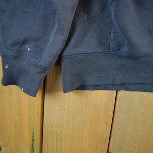K&ouml;nnte beinhalten: Nahaufnahme eines marineblauen Sweatshirt-&Auml;rmels und -Saums. Der Stoff weist Gebrauchsspuren auf, mit kleinen Farbspritzern am B&uuml;ndchen. Das Sweatshirt besteht aus einem dicken, strapazierf&auml;higen Material.