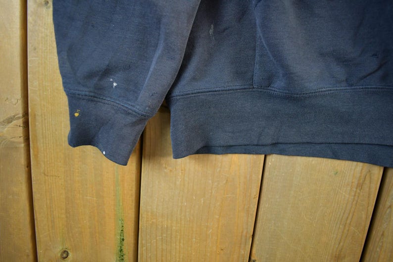 K&ouml;nnte beinhalten: Nahaufnahme eines marineblauen Sweatshirt-&Auml;rmels und -Saums. Der Stoff weist Gebrauchsspuren auf, mit kleinen Farbspritzern am B&uuml;ndchen. Das Sweatshirt besteht aus einem dicken, strapazierf&auml;higen Material.