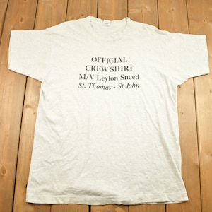 Może przedstawiać: Jasnoszary t-shirt z nadrukiem "OFFICIAL CREW SHIRT M/V Leylon Sneed St. Thomas - St. John" na przodzie.