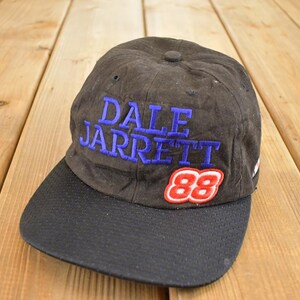 Peut inclure: Casquette de baseball noire avec une visière incurvée. Le chapeau porte le nom "DALE JARRETT" en lettres bleues et le numéro "88" en rouge et blanc. Le chapeau est probablement vintage.