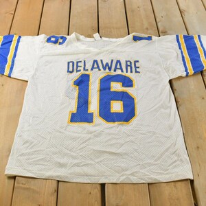 Peut inclure: Maillot de sport blanc en mesh avec des détails bleus et jaunes. Le mot "DELAWARE" est inscrit en bleu sur la poitrine, avec le numéro "16" en dessous, en bleu avec un contour jaune. Rayures bleues et jaunes sur les manches.