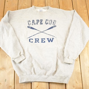 Puede incluir: Una sudadera gris claro con las palabras "CAPE COD CREW" en azul, con remos cruzados. La sudadera está colocada sobre una superficie de madera.
