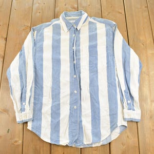 May include: A long-sleeved button-down shirt featuring a vertical striped pattern in light blue and white. The shirt has a standard collar, a single chest pocket on the left side, and white buttons running down the centre front placket and on the cuffs.