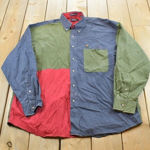 May include: A long-sleeve button-down shirt featuring a color-blocked design in shades of blue, olive green, and red. The shirt includes a button-down collar, a single chest pocket, and buttoned cuffs. The interior collar lining is red.