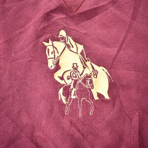 May include: A maroon t-shirt with a gold silhouette of two people riding horses.