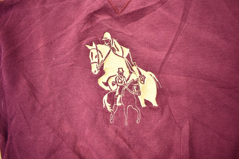 May include: A maroon t-shirt with a gold silhouette of two people riding horses.