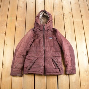 May include: A maroon quilted jacket with a fleece-lined hood and a zipper closure. The jacket has a small logo patch on the left chest.
