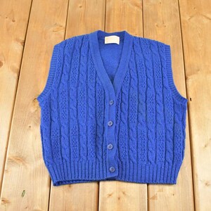 May include: A vibrant royal blue knit sweater vest featuring a classic cable knit pattern, a V-neckline, and a front button closure with four matching blue buttons. The garment has ribbed trim along the armholes, the neckline, and the bottom hem.