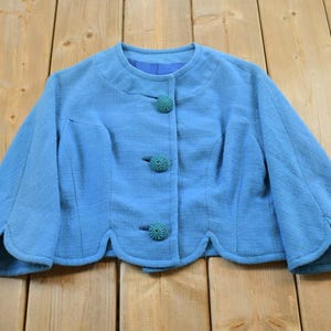 Puede incluir: Chaqueta azul claro vintage con cuello redondo y tres botones a juego. La chaqueta tiene un dobladillo festoneado único y mangas tres cuartos. La tela tiene una apariencia texturizada, y la chaqueta está colocada sobre una superficie de madera.
