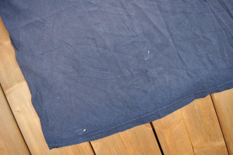 May include: A dark blue t-shirt with a smooth, slightly textured surface. The t-shirt is folded and lying on a wooden surface.