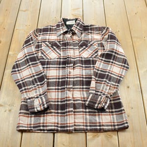 May include: A long-sleeve button-up shirt featuring a plaid pattern in shades of dark brown, cream, and rust orange. The shirt includes a standard collar, two chest pockets with buttoned flaps, and buttoned cuffs, showcasing a structured design.