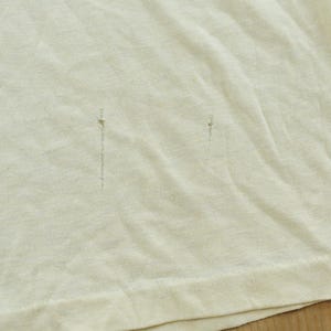 May include: A plain off-white t-shirt featuring two small, vertical, dark-colored pinhole-style imperfections or snags located in the center of the fabric, with a visible hemline along the bottom edge of the garment.
