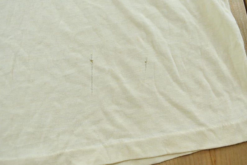 May include: A plain off-white t-shirt featuring two small, vertical, dark-colored pinhole-style imperfections or snags located in the center of the fabric, with a visible hemline along the bottom edge of the garment.