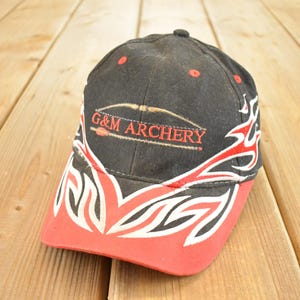 Könnte beinhalten: Schwarze Baseballkappe mit rotem Schirm und Akzenten. Auf der Kappe steht der Text "G&M ARCHERY" in Rot, zusammen mit einer Pfeil- und Bogen-Grafik. Weiße und rote flammenartige Designs zieren die Seiten der Kappe.