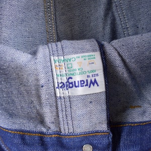 Op de afbeelding: Een wit Wrangler-label van denim in maat 18 met de tekst "Wrangler", "SIZE 18", "100% COTTON/CDTION", "CA 00121", "MADE IN", "FABRIQUE AU CANADA".