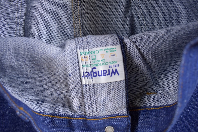 Op de afbeelding: Een wit Wrangler-label van denim in maat 18 met de tekst "Wrangler", "SIZE 18", "100% COTTON/CDTION", "CA 00121", "MADE IN", "FABRIQUE AU CANADA".