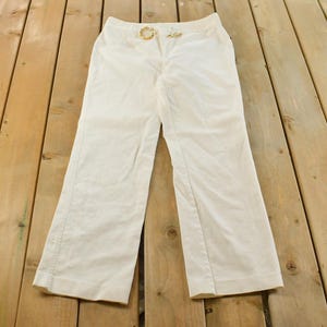 May include: Cream-colored pants with a gold-tone buckle belt. The pants have a straight leg design and are made of a lightweight fabric. The pants are laid flat on a wooden surface.