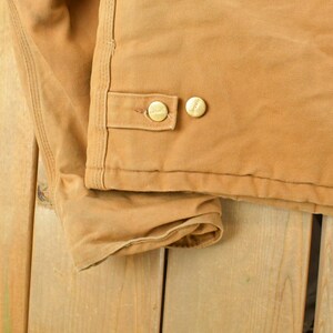 Puede incluir: Primer plano de una chaqueta de trabajo color canela. La chaqueta presenta dos botones dorados en una solapa y est&aacute; hecha de una tela duradera y resistente. La chaqueta est&aacute; sobre una superficie de madera.