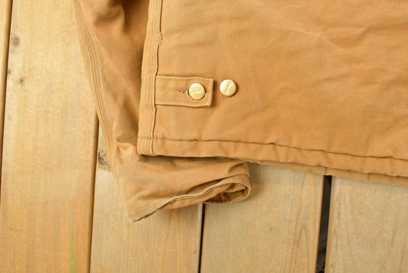 Puede incluir: Primer plano de una chaqueta de trabajo color canela. La chaqueta presenta dos botones dorados en una solapa y est&aacute; hecha de una tela duradera y resistente. La chaqueta est&aacute; sobre una superficie de madera.