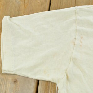 May include: A short-sleeved, off-white t-shirt featuring a crew neckline and visible light brown staining on the fabric near the shoulder seam and sleeve area. The garment is constructed with standard stitching along the sleeve hem and shoulder.