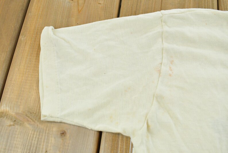 May include: A short-sleeved, off-white t-shirt featuring a crew neckline and visible light brown staining on the fabric near the shoulder seam and sleeve area. The garment is constructed with standard stitching along the sleeve hem and shoulder.