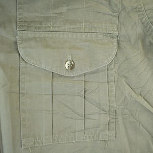 Peut inclure: Gros plan d'une chemise vert olive clair avec une poche sur la poitrine. La chemise a un design boutonn&eacute; avec deux boutons visibles. Le tissu semble l&eacute;g&egrave;rement froiss&eacute;.