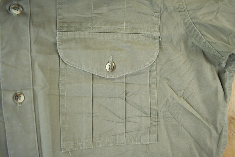 Peut inclure: Gros plan d'une chemise vert olive clair avec une poche sur la poitrine. La chemise a un design boutonn&eacute; avec deux boutons visibles. Le tissu semble l&eacute;g&egrave;rement froiss&eacute;.