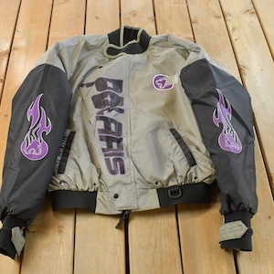 May include: A vintage tan and black jacket with the word "Polaris" printed in large, dark letters. The jacket has black sleeves with purple flame designs and a purple logo on the chest. The jacket has a black collar and cuffs.