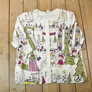 Puede incluir: Un cárdigan blanco de manga larga con un estampado navideño de personas comprando y árboles. El estampado incluye las palabras "Shop in City" y "Plaisance".