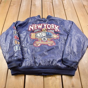May include: A blue satin baseball jacket with the words "New York Division Champions" and a baseball-themed graphic on the back. The jacket has a ribbed collar, cuffs, and waistband.