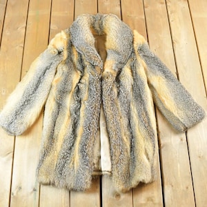 May include: A vintage fur coat with a collar, displayed on a wooden surface. The coat features a mix of gray, tan, and cream colors. The coat is open, revealing the lining. The fur appears soft and plush.