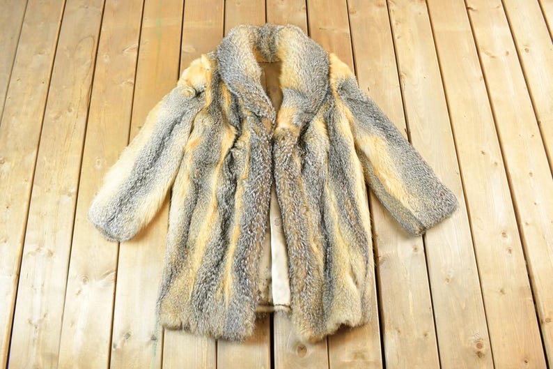 May include: A vintage fur coat with a collar, displayed on a wooden surface. The coat features a mix of gray, tan, and cream colors. The coat is open, revealing the lining. The fur appears soft and plush.