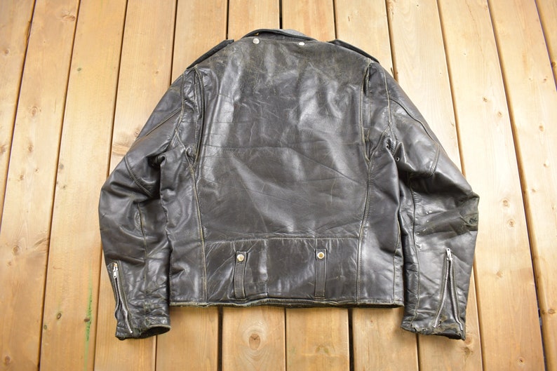 Puede incluir: Una chaqueta de cuero negra para motocicleta con cierre de cremallera y cuello con bot&oacute;n a presi&oacute;n. La chaqueta tiene un aspecto desgastado y est&aacute; usada.