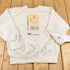 May include: A light gray Champion sweatshirt featuring the text "Vanderbilt" and "Final Four Atlanta 1995". The design includes a graphic of an apple and the Champion logo in purple and red. The sweatshirt has long sleeves.