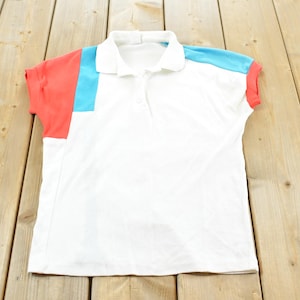 Puede incluir: Polo blanco con mangas cortas. La camisa presenta detalles de bloques de color coral y azul claro en las mangas y los hombros. Fabricada con un material suave y ligero.