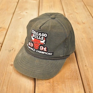 Op de afbeelding: Olijfgroene Chicago Bulls baseballpet met een gebogen klep. De pet heeft een patch met het teamlogo, het jaar 1991 en de woorden "World Champions". De pet heeft een touwdetail rond de klep.
