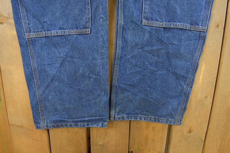 K&ouml;nnte beinhalten: Nahaufnahme einer blauen Jeans-Arbeitshose. Die Hose hat gro&szlig;e quadratische Taschen auf der Vorderseite der Beine und besteht aus einem strapazierf&auml;higen Denim-Stoff. Die N&auml;hte sind sichtbar, und die Hose scheint abgenutzt zu sein.