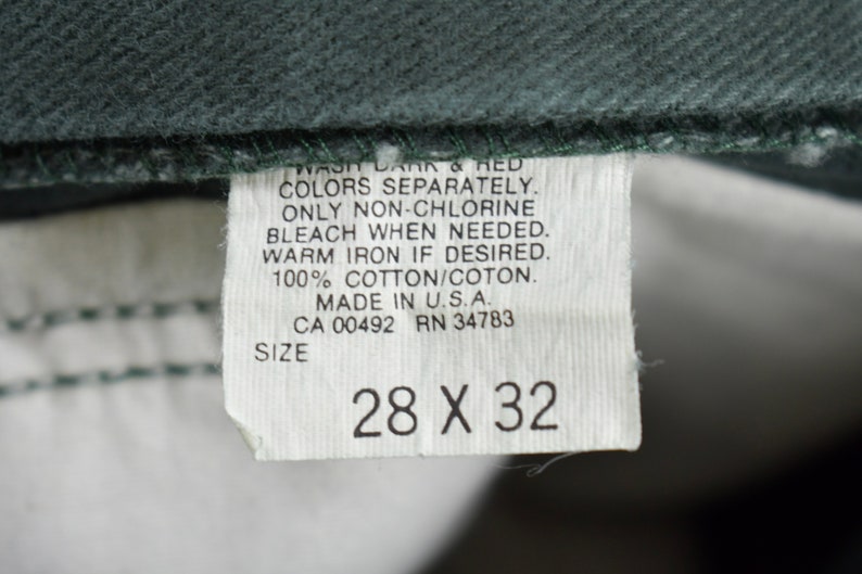 Może przedstawiać: Biała metka z czarnym tekstem, kt&oacute;ry brzmi: "WASH DARK & RED COLORS SEPARATELY. ONLY NON-CHLORINE BLEACH WHEN NEEDED. WARM IRON IF DESIRED. 100% COTTON/COTON. MADE IN U.S.A. CA 00492 RN 34783 SIZE 28 X 32"