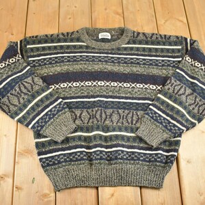 May include: A vintage crew neck sweater in shades of green, blue, and white. The sweater features a geometric pattern with horizontal stripes and repeating shapes. The long sleeves and body are made of a knitted material.