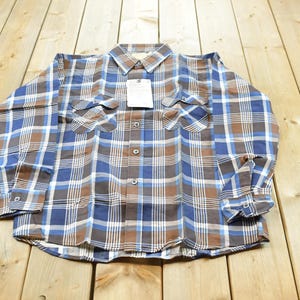 May include: A long-sleeve button-down shirt featuring a plaid pattern in shades of brown, blue, and white. The shirt includes a standard collar, two chest pockets with buttoned flaps, and a central button placket with a white rectangular tag attached.