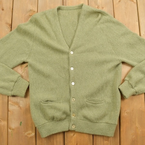 May include: A green knitted cardigan with a v-neck and button closure. The cardigan has two pockets and a ribbed hem.