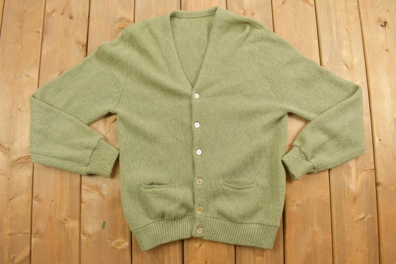 May include: A green knitted cardigan with a v-neck and button closure. The cardigan has two pockets and a ribbed hem.