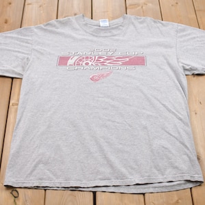May include: A gray t-shirt with a red and white graphic that says "2002 Stanley Cup Champions" with a red winged wheel logo.