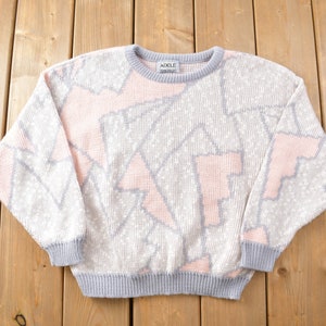 May include: A pink and grey sweater with a geometric pattern. The sweater has a crew neck and long sleeves. The label on the sweater reads "ADILE".