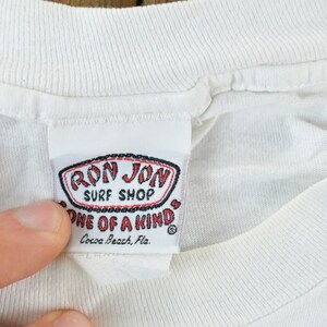Puede incluir: Primer plano de una etiqueta de camiseta blanca de Ron Jon Surf Shop. La etiqueta es rectangular con un logotipo rojo y negro que dice "Ron Jon Surf Shop" y "One of a Kind". La etiqueta tambi&eacute;n incluye la ubicaci&oacute;n "Cocoa Beach, Fla."